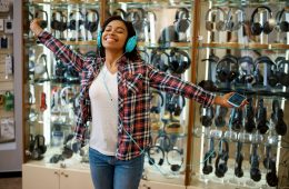 Happy customer choosing headphones in audio store, music fan. Female person in music shop, showcase with earphones on background, buyer in multimedia salon